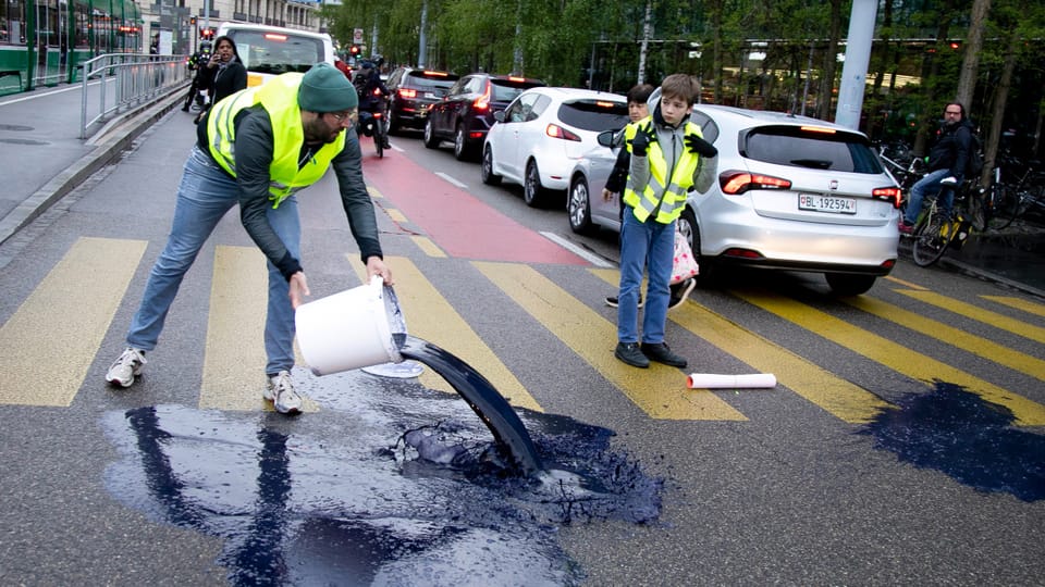 Basel: Klimaaktivisten blockieren Morgenverkehr und leeren Flüssigkeit auf Strasse.